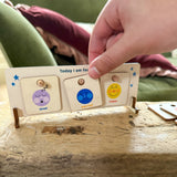 Hand placing "worried" token on a wooden feelings chart next to "tired" and "happy"