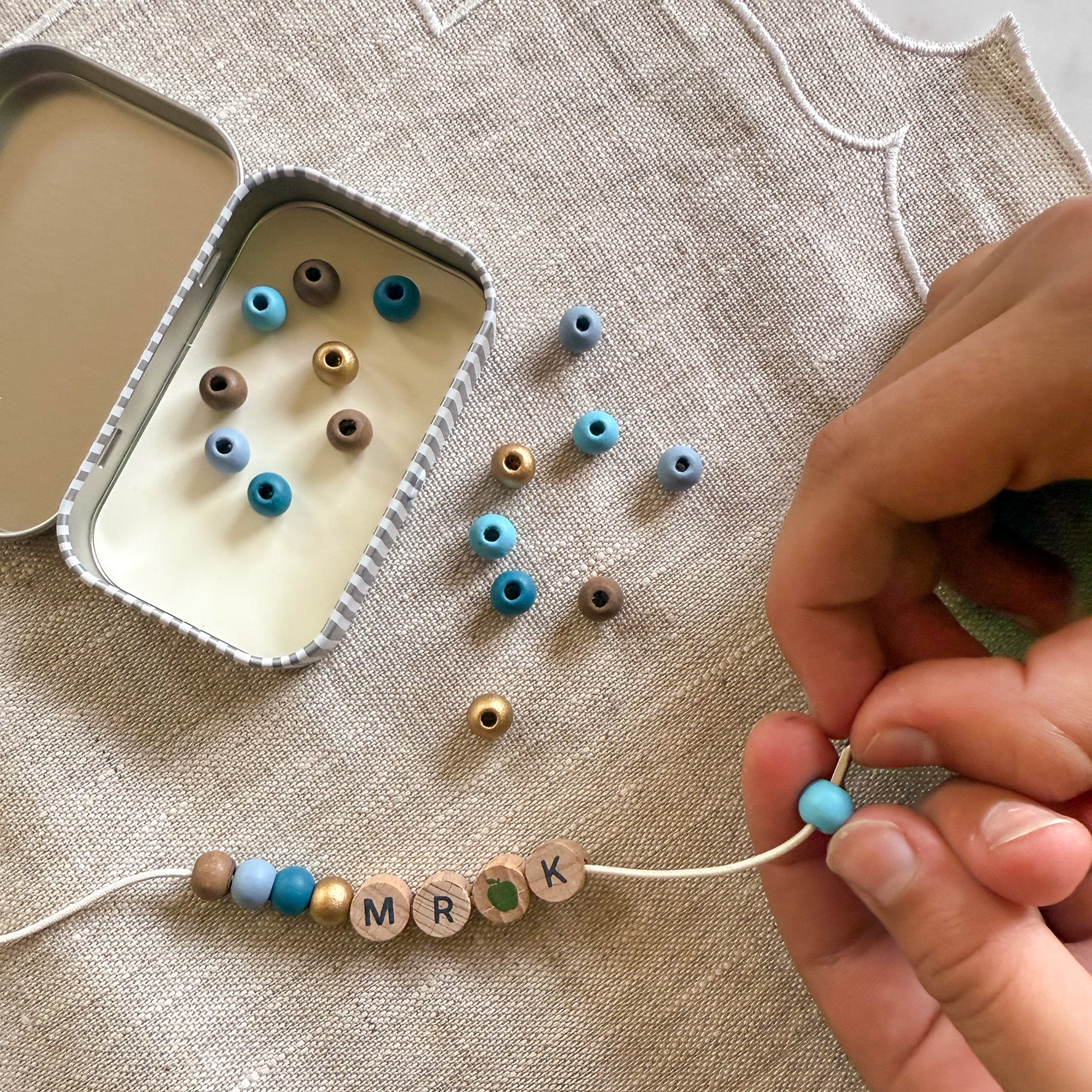 Close-up of hands threading blue beads onto a bracelet with "MR K" spelled out, next to an open tin.