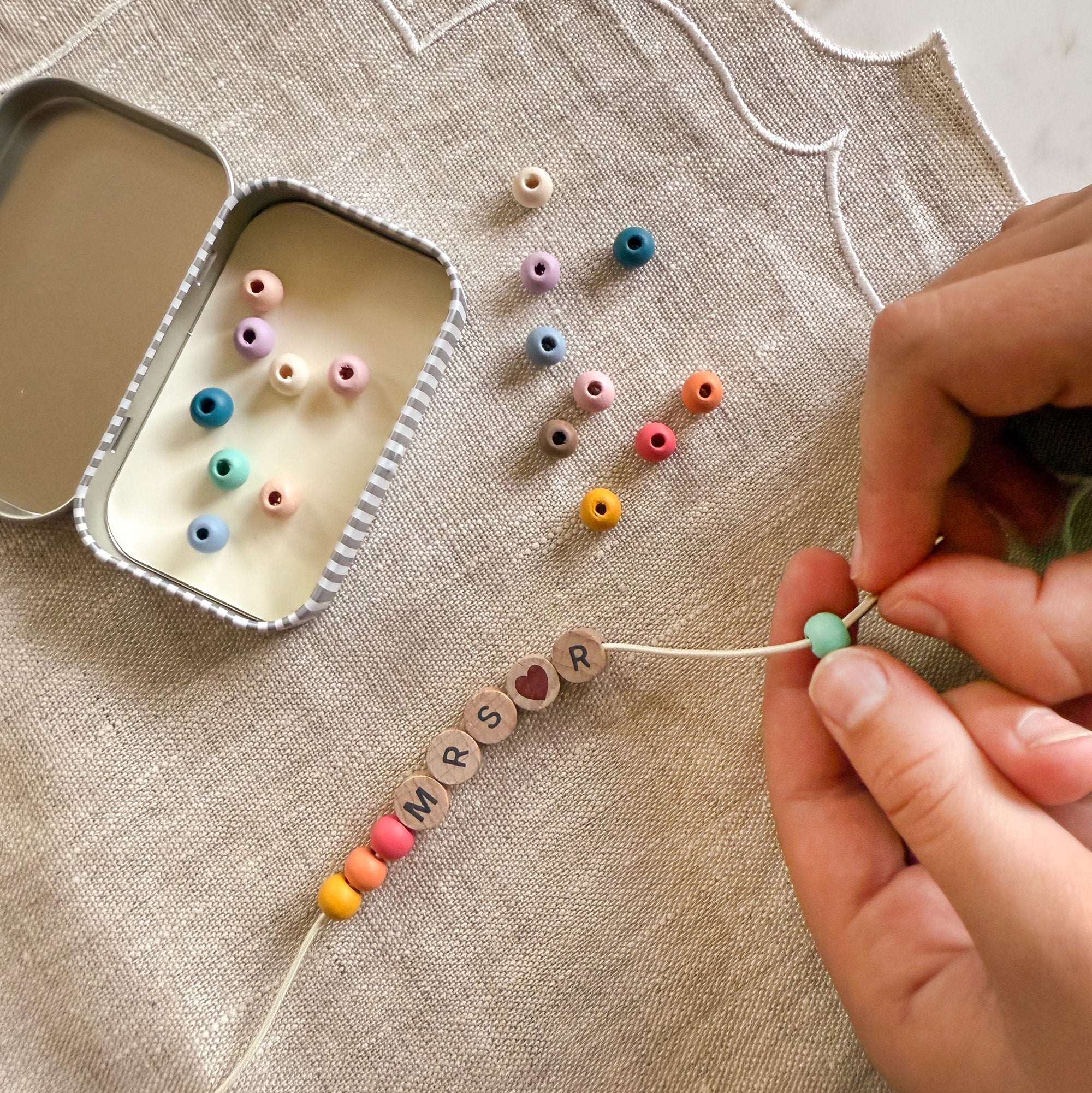 Close-up of hands threading rainbow coloured beads onto a bracelet with "MRS R" spelled out, next to an open tin.