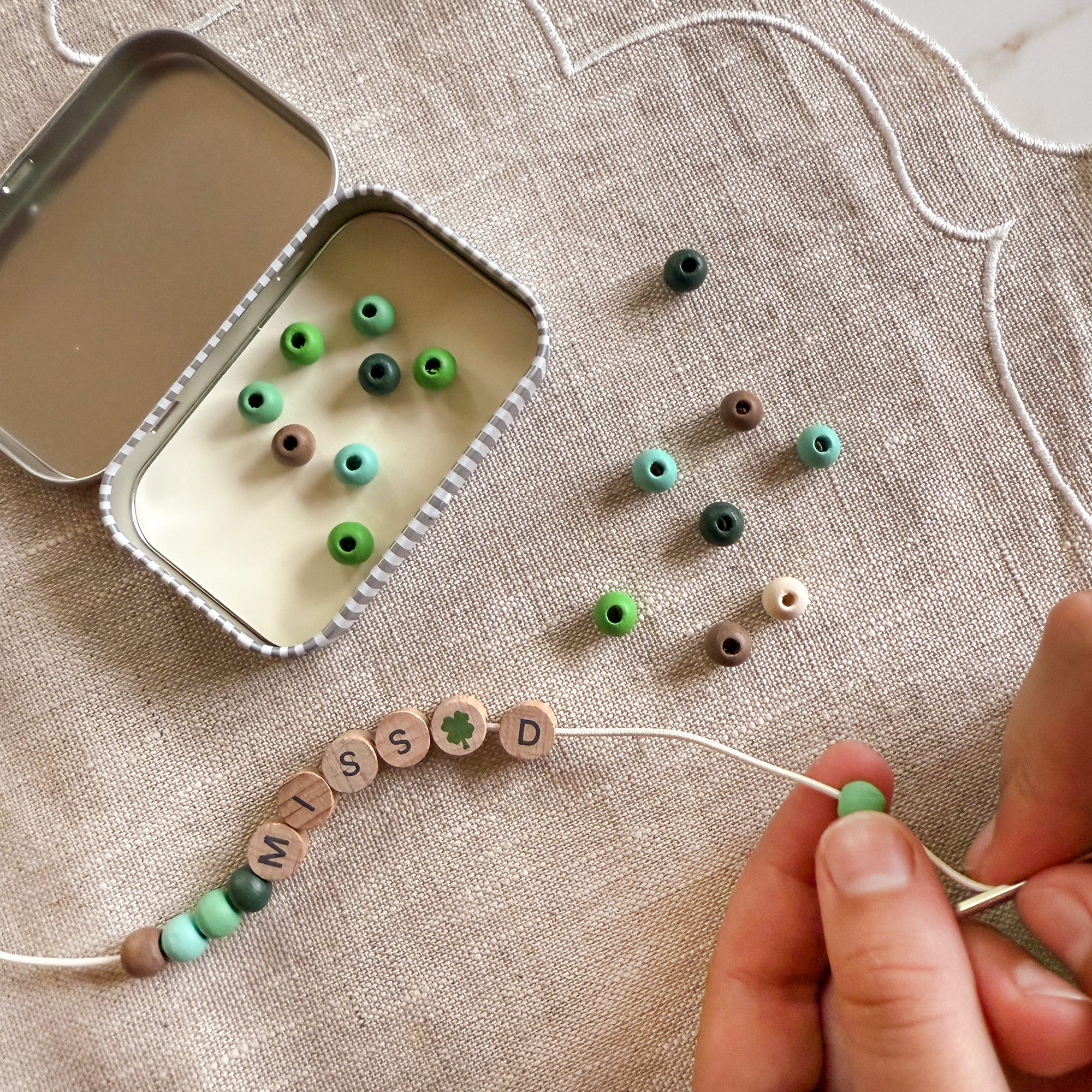 Close-up of hands threading green beads onto a bracelet with "MISS D" spelled out, next to an open tin.