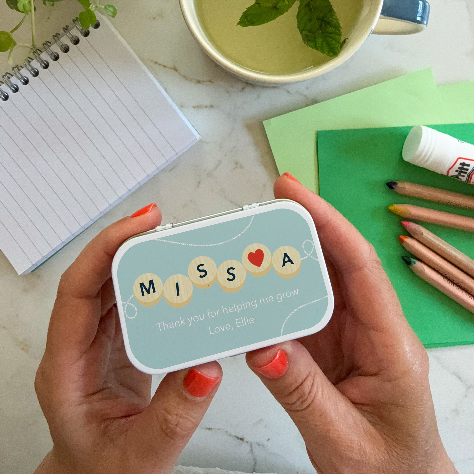 Hands holding a personalised tin with wooden bead design spelling "MISS A" and message: "Thank you for helping me grow."