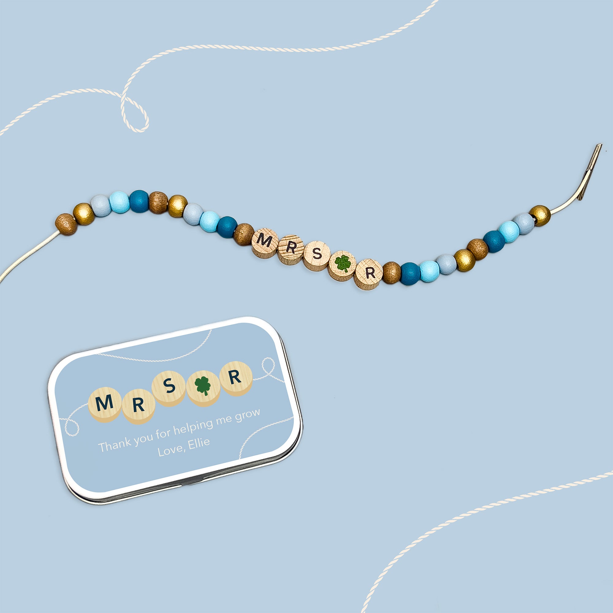 Bracelet with "MRS R" and green clover bead beside a matching personalised tin on a blue background.