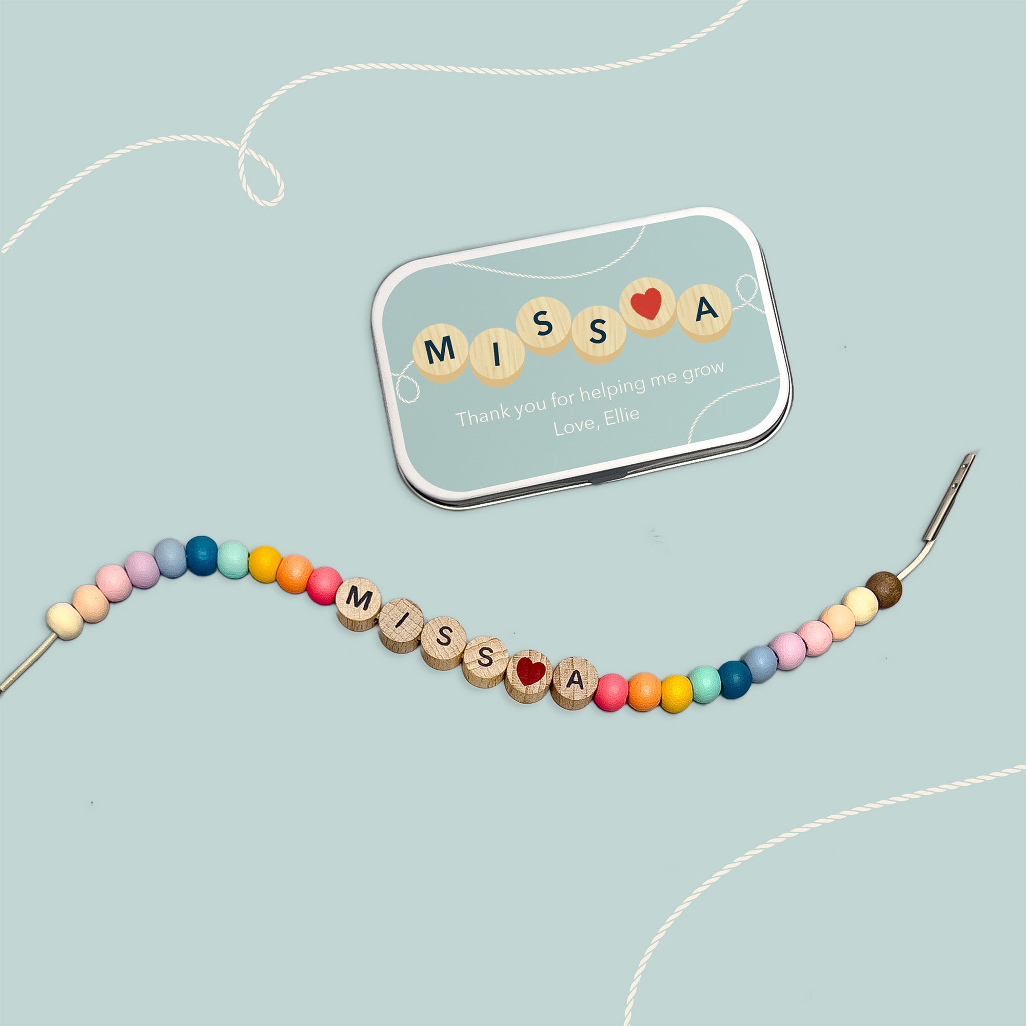 Bracelet with "MISS A" and red heart bead beside a matching personalised tin on a pale blue background.