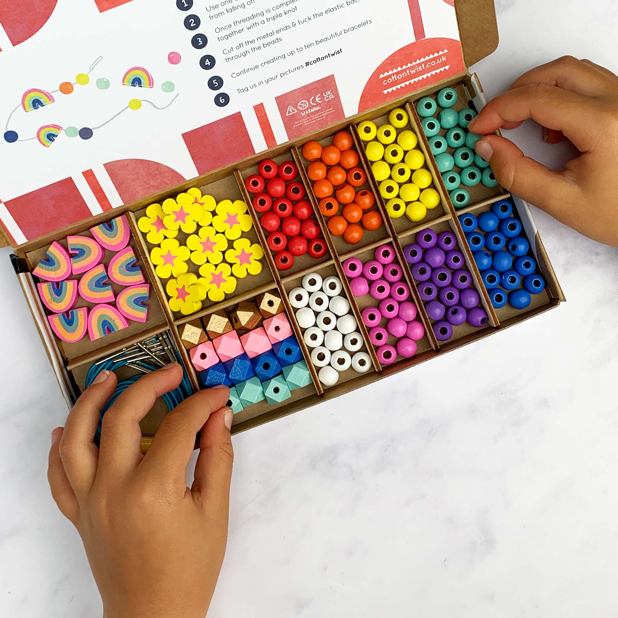 Hands selecting wooden beads from a colourful beading kit box with rainbow and flower shapes.