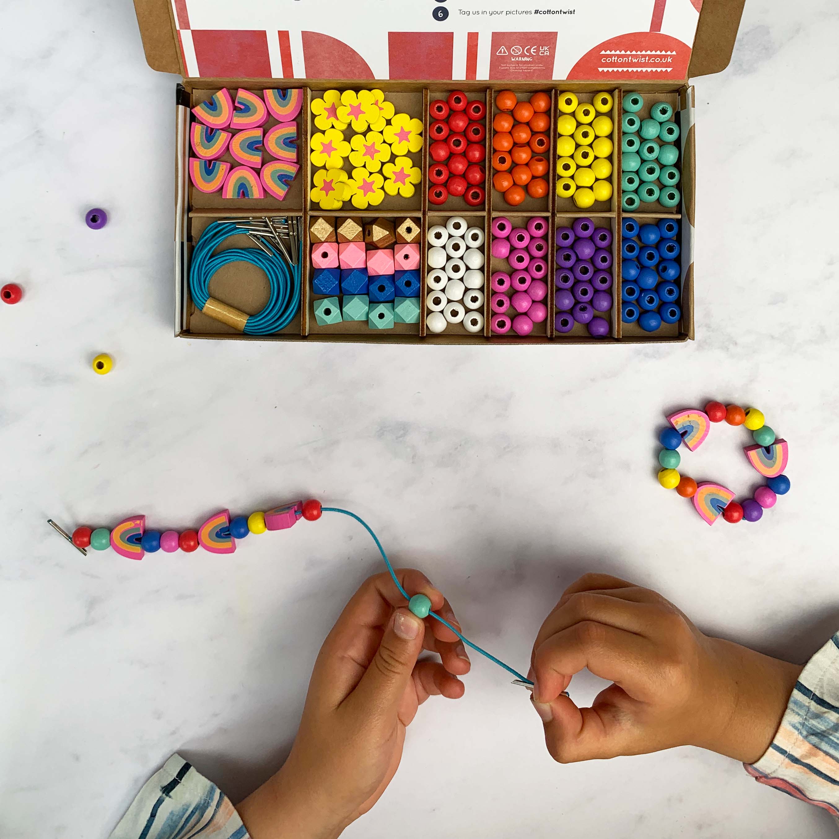 Child threading beads to finish a rainbow bracelet, with full kit open above and a completed bracelet nearby.