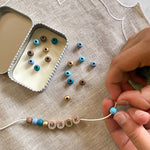 Close-up of hands threading blue beads onto a bracelet with "MR K" spelled out, next to an open tin.