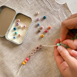 Close-up of hands threading rainbow coloured beads onto a bracelet with "MRS R" spelled out, next to an open tin.