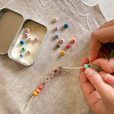 Close-up of hands threading rainbow coloured beads onto a bracelet with "MRS R" spelled out, next to an open tin.