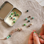 Close-up of hands threading green beads onto a bracelet with "MISS D" spelled out, next to an open tin.