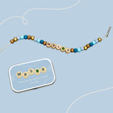 Bracelet with "MRS R" and green clover bead beside a matching personalised tin on a blue background.