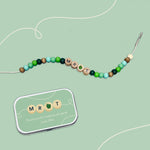 Bracelet with "MR T" and green apple bead beside a matching personalised tin on a green background.