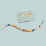 Bracelet with "MISS A" and red heart bead beside a matching personalised tin on a pale blue background.